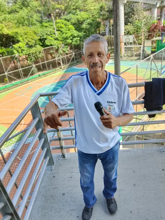 Jaime Ramirez con camiseta blanca bordada con su nombre, «Jimmy», sosteniendo un micrófono sobre una cancha comunitaria